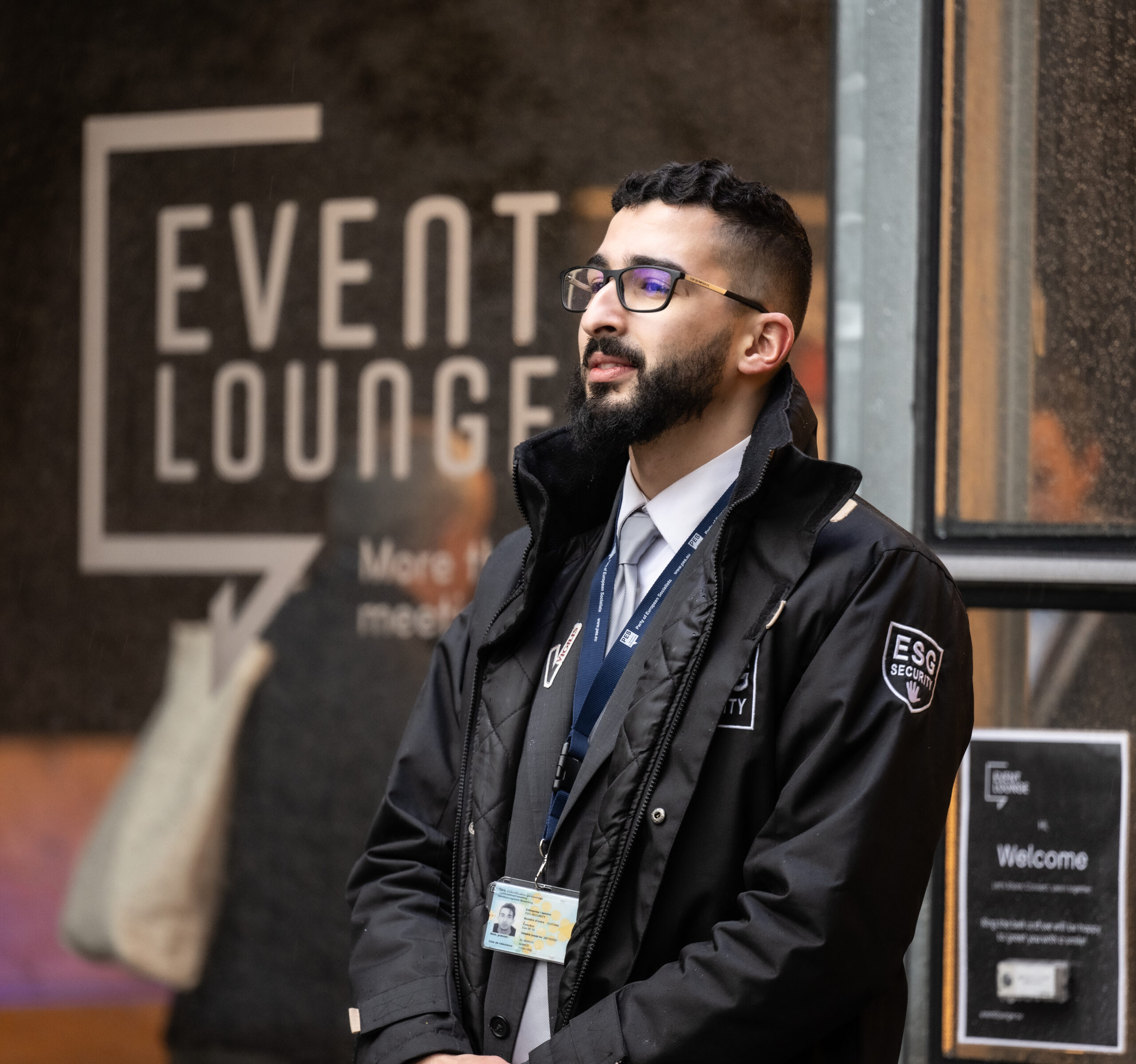 Agent de sécurité ESG sur le site d’un événement, portant une veste noire et badge, devant une affiche “Event Lounge”.